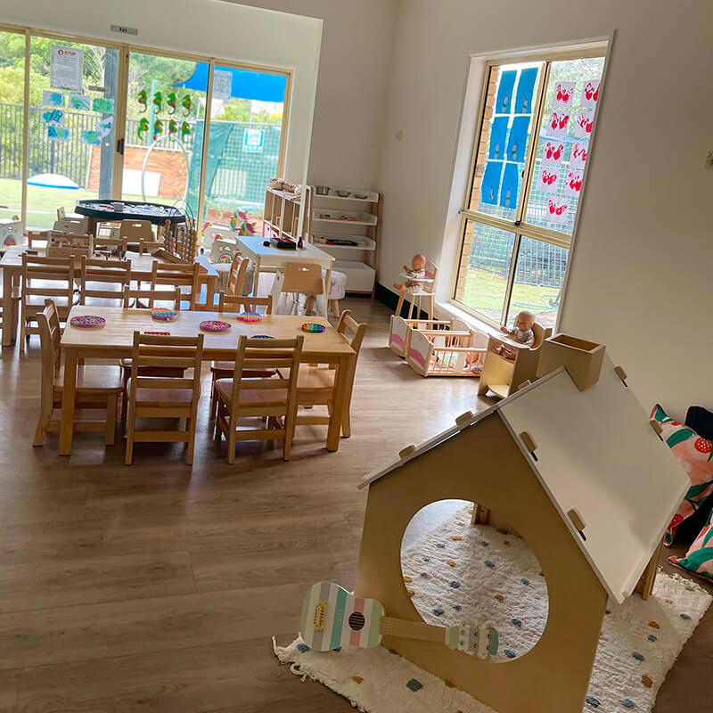A brightly lit kindergarten playroom with wooden furniture, small tables, and chairs. Toys are scattered around, including a white playhouse, dolls, and art supplies. Large windows provide natural light and a view of a green outdoor area, making it perfect for day care or before school care activities.