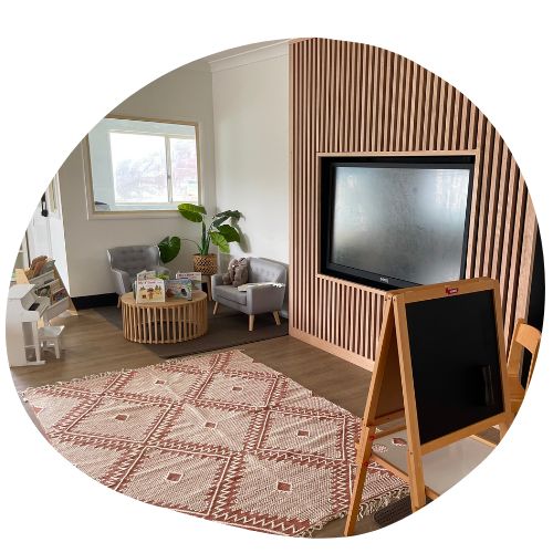 A cozy children's playroom designed for afterschool care, with a TV mounted on a wall adorned with vertical wooden slats. It features a small armchair, a round wooden table, a patterned rug, a blackboard easel, and a bookshelf. A window lets in natural light, and a potted plant adds greenery.
