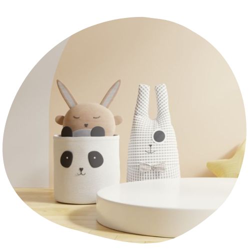 A cozy room with light-colored walls features a stuffed bunny peeking out of a basket with a panda face illustration. Nearby, a plush bunny with a checkered pattern and large eyes sits on the floor next to a white round table, creating an inviting space for before school care in kindergarten.